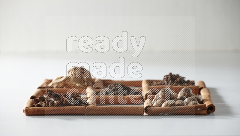 9 مربعات من أعواد القرفة ممتلئة بالشاي في المنتصف محاطة بجوزة الطيب والنعناع المجفف والقرنفل والريحان المجفف والزنجبيل المجفف والقرفة واليانسون والهيل على أرضية بيضاء