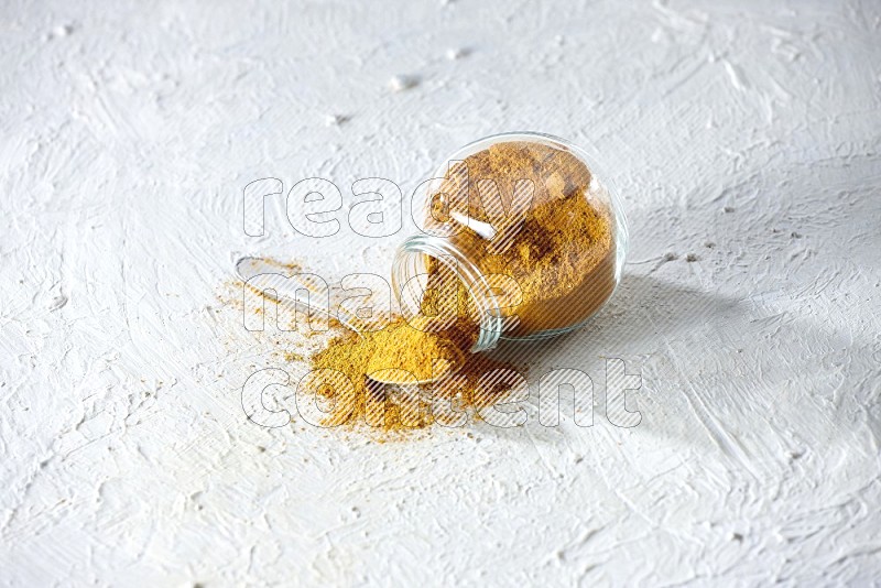 A flipped glass spice jar and a metal spoon full of turmeric powder and powder spilled out of it on textured white flooring