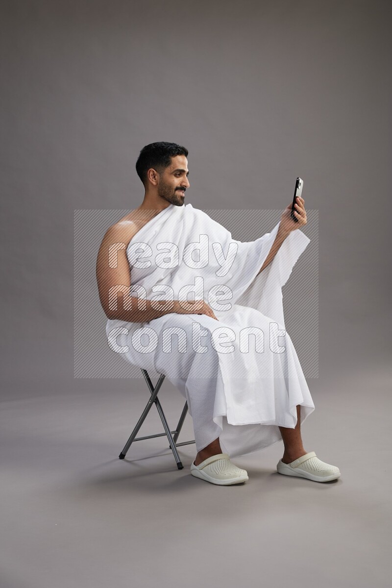 A man wearing Ehram sitting on chair taking selfie on gray background