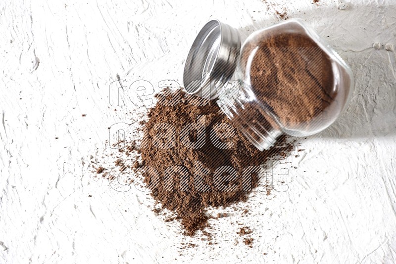 A flipped glass spice jar full of cloves powder and powder came out of it on textured white flooring