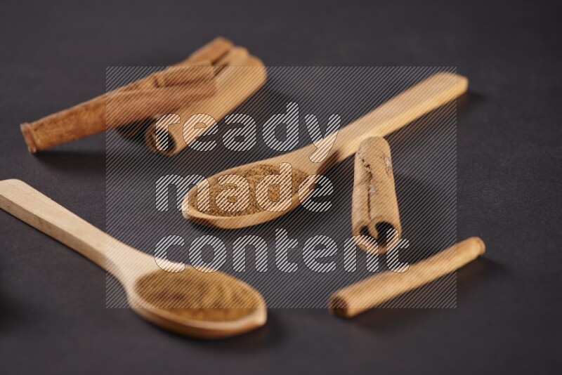 Two wooden spoons full of cinnamon powder with cinnamon sticks on black background