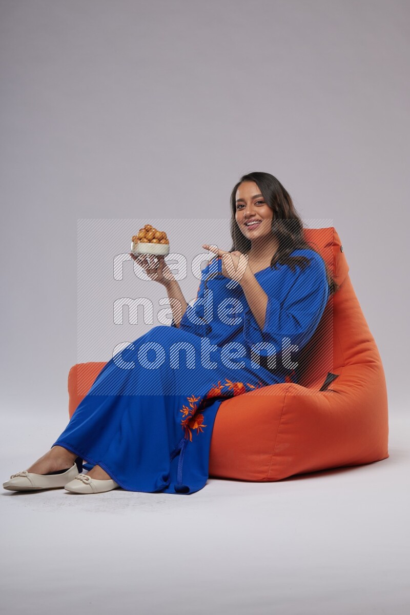 A Woman sitting on an orange beanbag wearing Jalabeya holding a plate of luqaimat