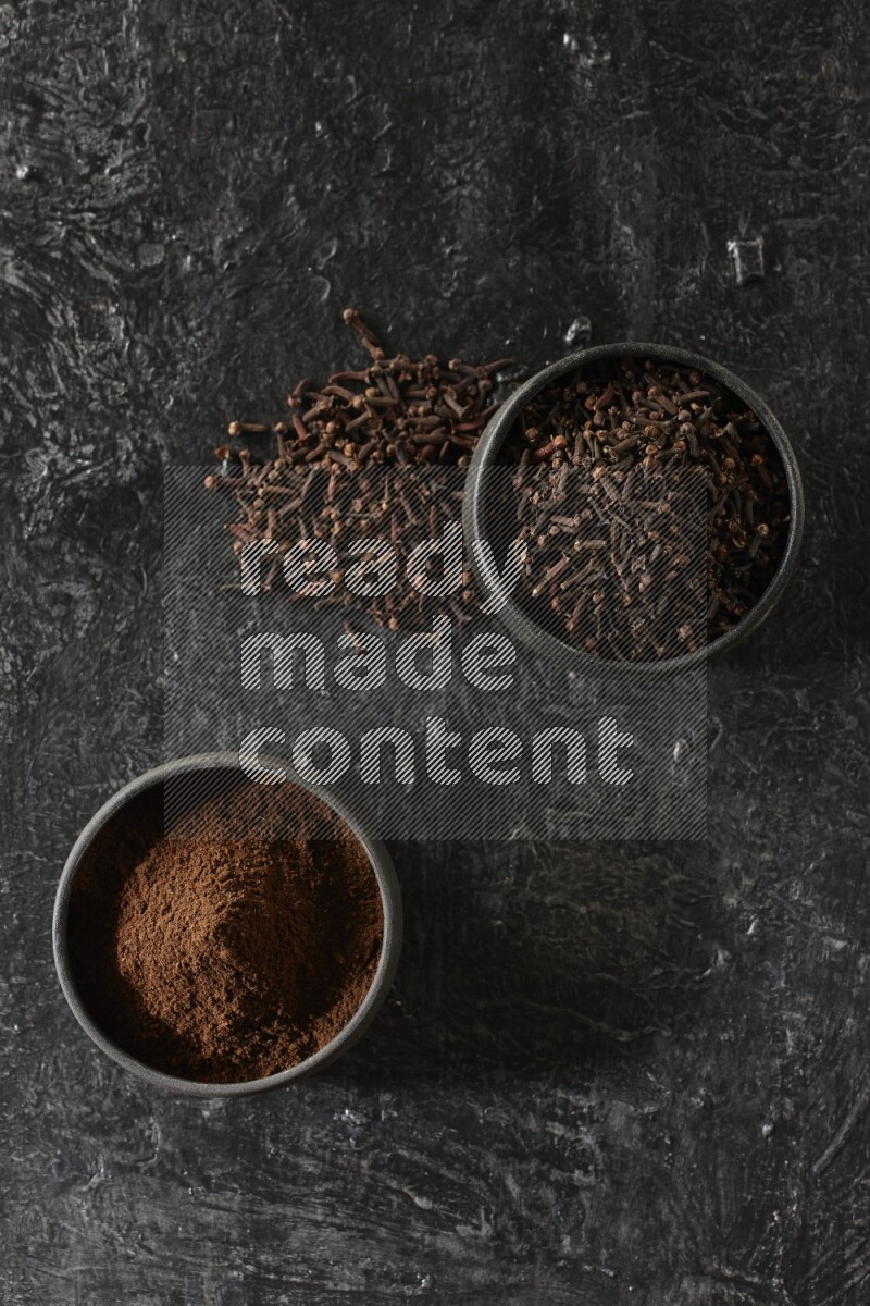 2 Black pottery bowls full of cloves and the other full of cloves powder on textured black flooring