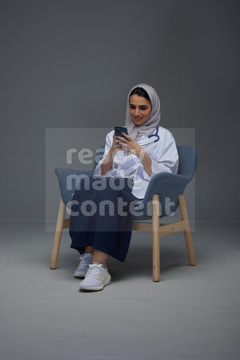 A doctor wearing a light gray head scarf sitting on a dark grey chair and using a laptop while talking in the phone eye level on a grey background