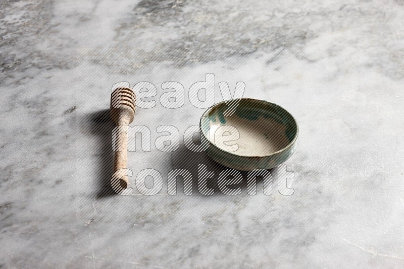 Multicolored Pottery bowl with wooden honey handle on the side with grey marble flooring, 45 degree angle