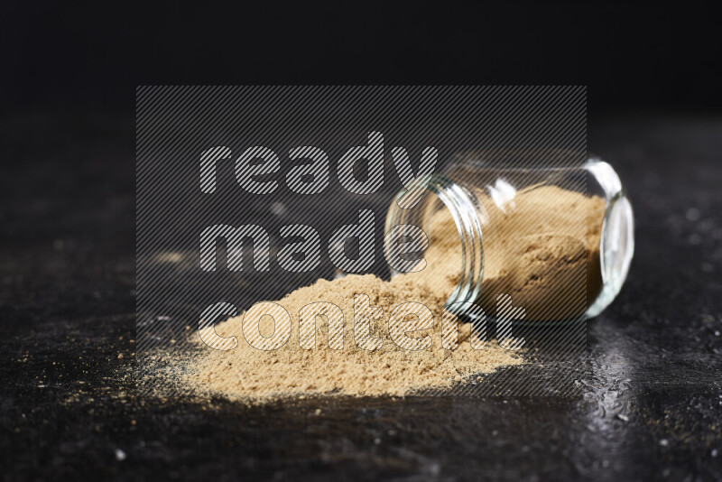 A glass jar full of ground ginger powder flipped with some spilling powder on black background