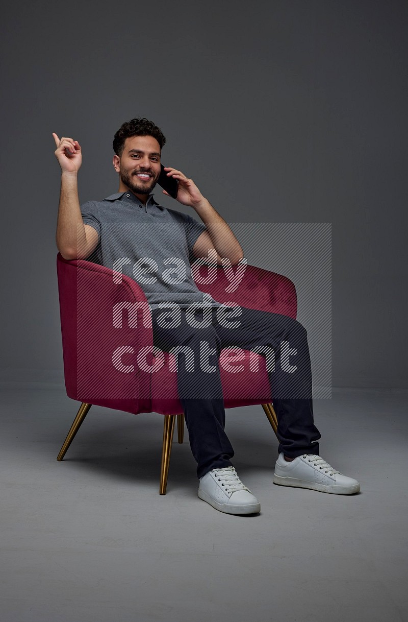 A man wearing casual talking in the phone and making multi hand gestures while sitting on a burgundy chair different angles eye level on a gray background