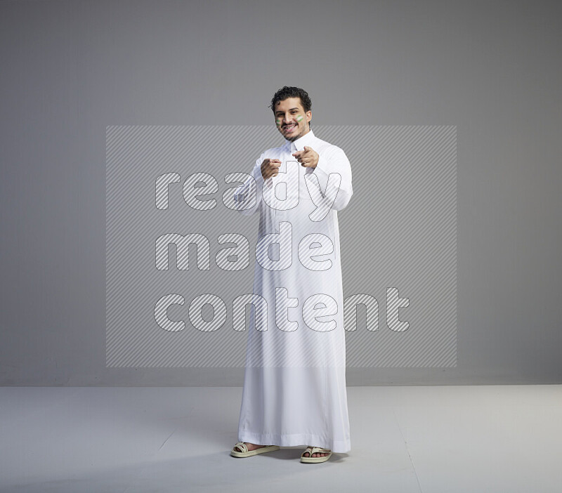 A Saudi man standing wearing thob with face painting interacting with the camera on gray background