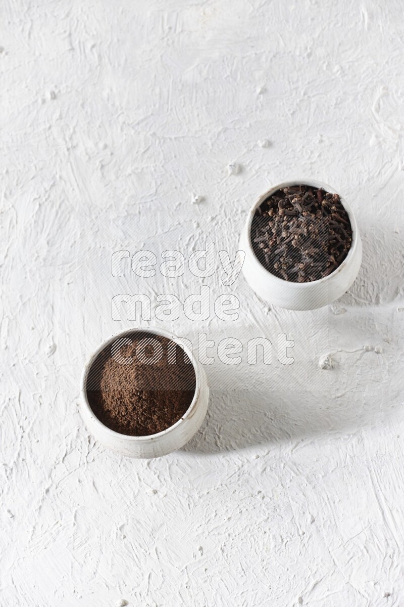 2 Beige ceramic bowls, one full of cloves and the other full of powder on textured white flooring