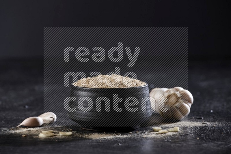 A black pottery bowl full of garlic powder with garlic cloves and bulb beside it on a textured black flooring in different angles