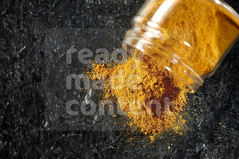 A flipped glass jar full of turmeric powder and powder spilled out of it on a textured black flooring