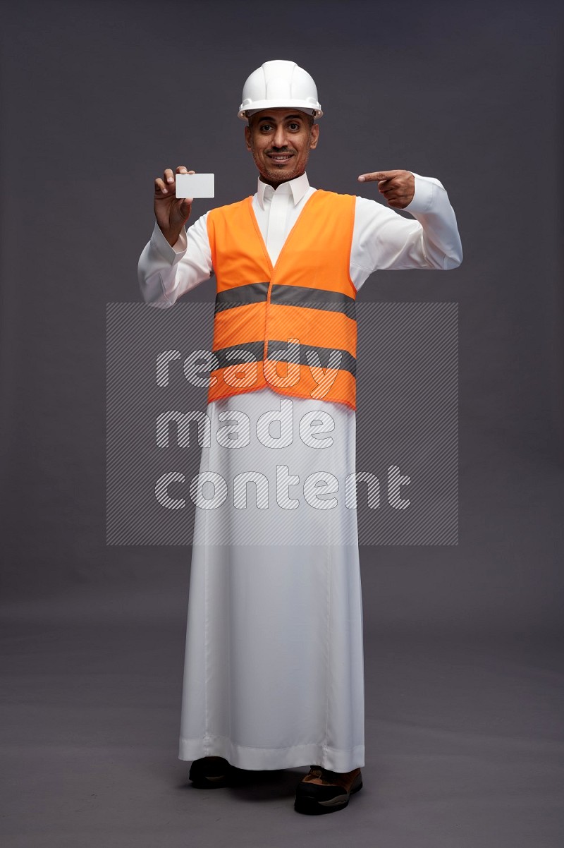 Saudi man wearing thob with engineer vest standing holding ATM card on gray background