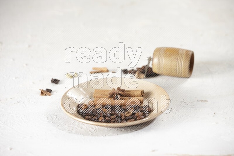 Beige plate full of coffee beans, cinnamon sticks and star anise with a coffee grinder, coffee beans, cinnamon pieces and cardamom next of it on white background
