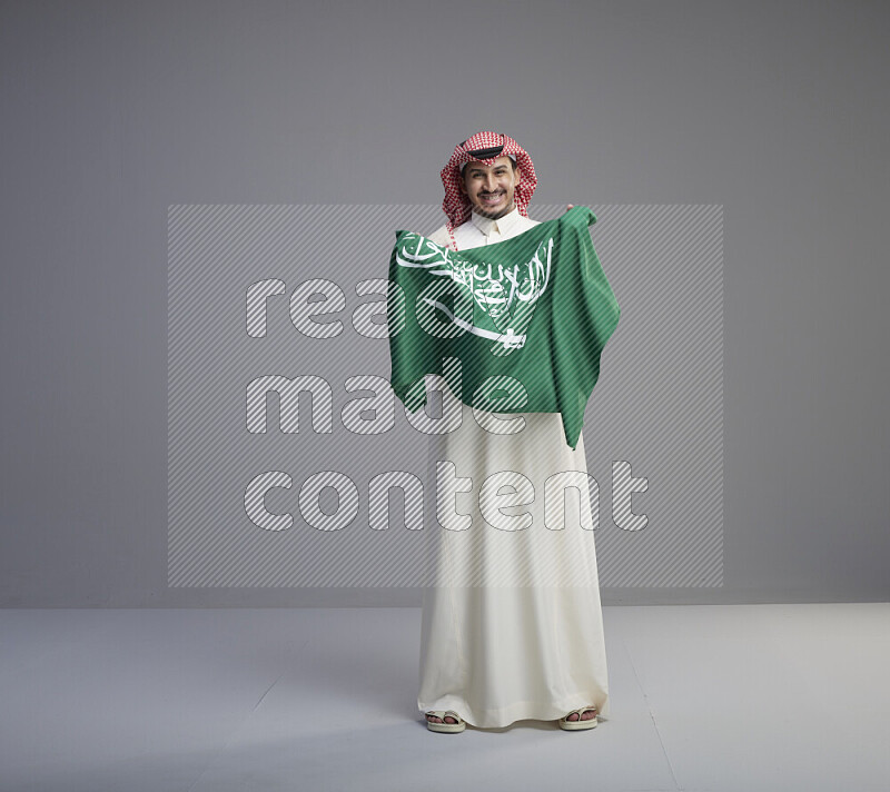 A saudi man standing wearing thob and red shomag holding big saudi flag on gray background