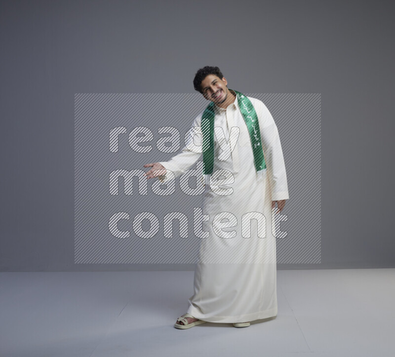 A Saudi man standing wearing thob and Saudi flag scarf on gray background
