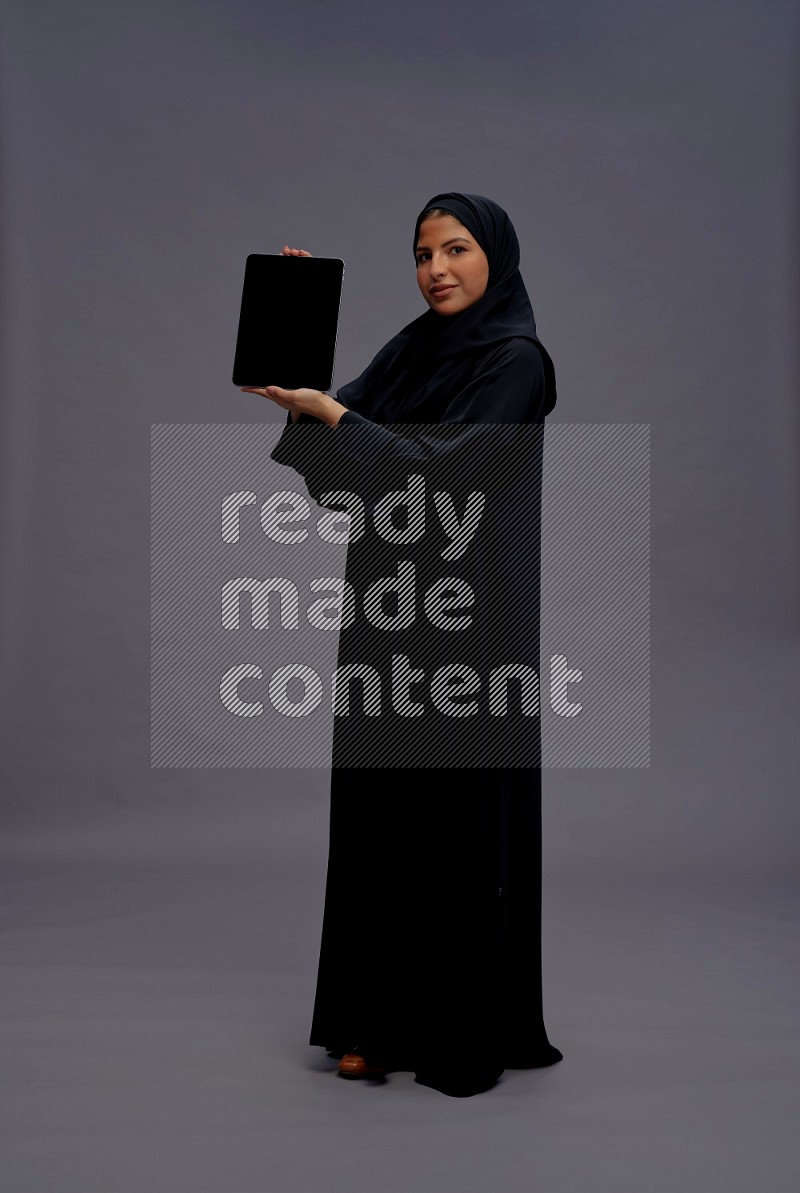 Saudi woman wearing Abaya standing showing tablet to camera on gray background