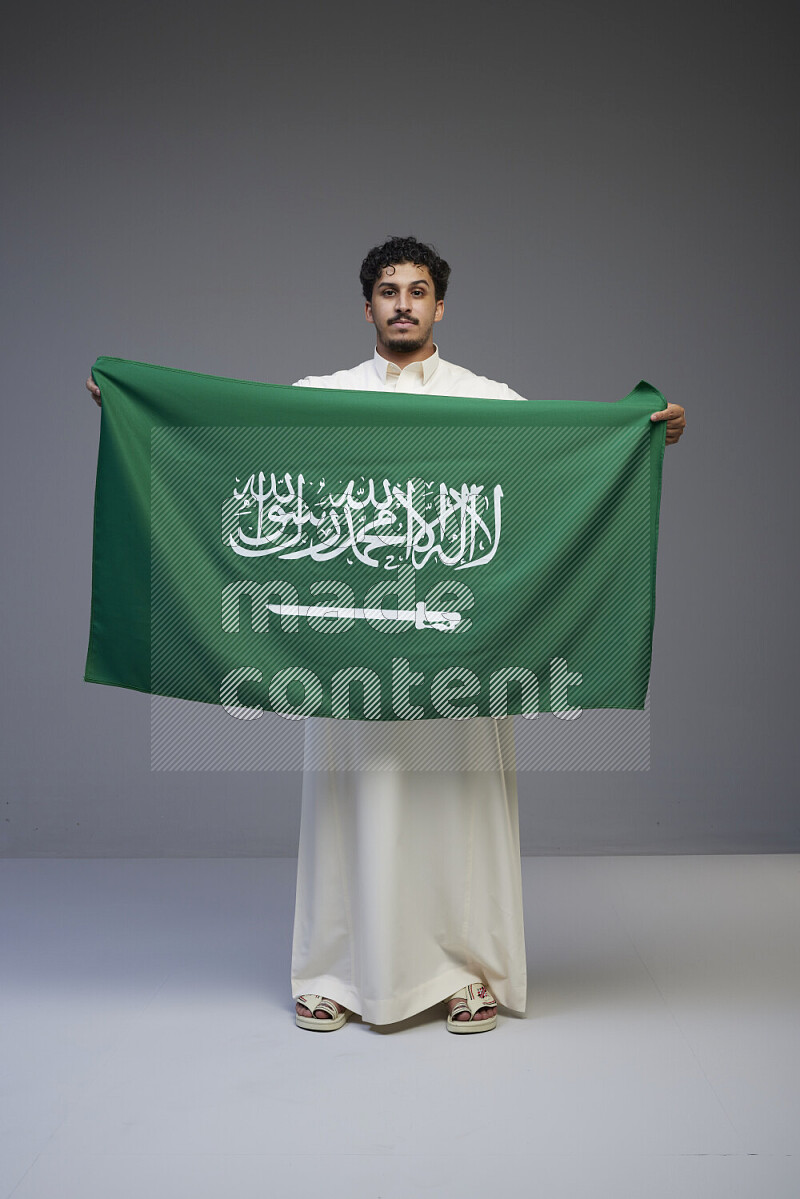 A Saudi man standing wearing thob holding big Saudi flag on gray background