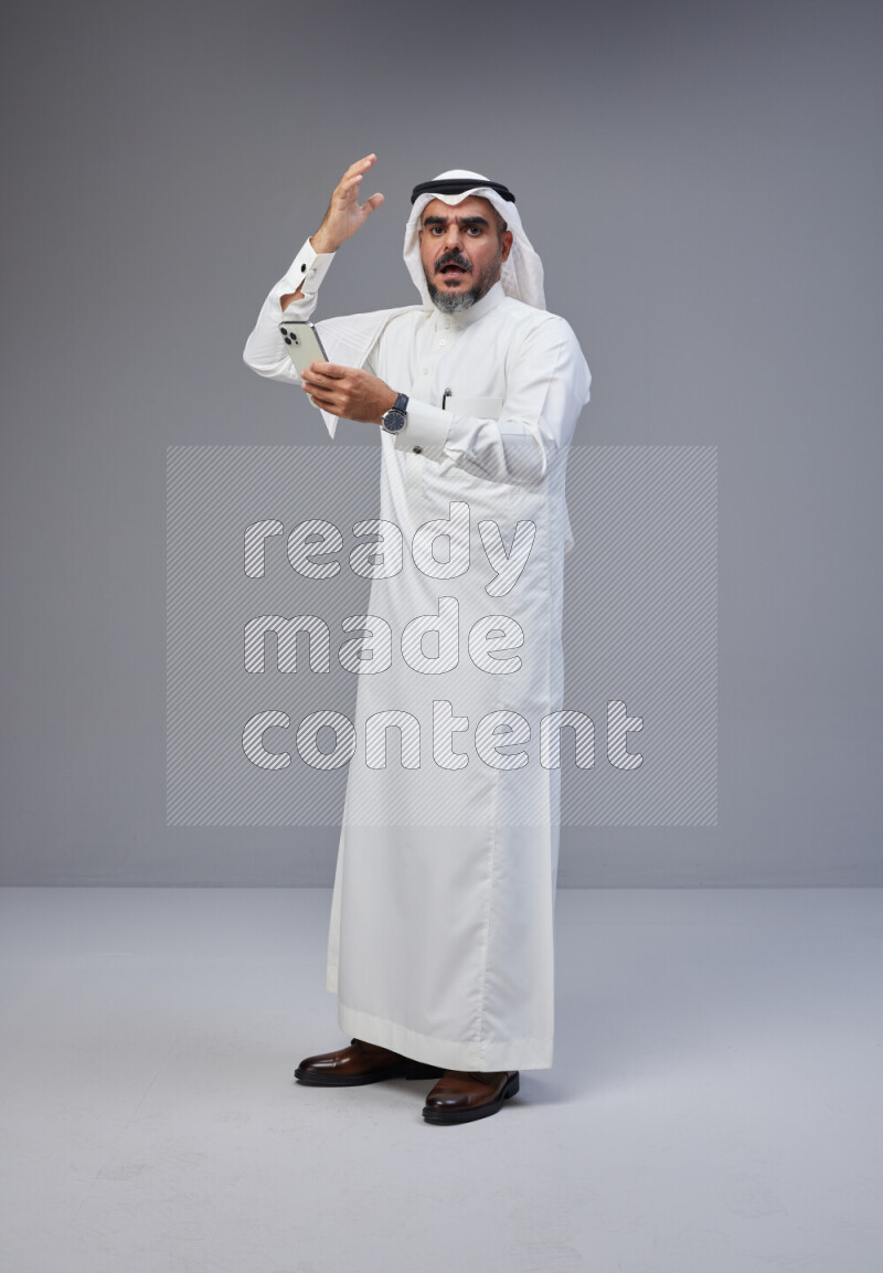 Saudi man Wearing Thob and white Shomag standing texting on phone on Gray background