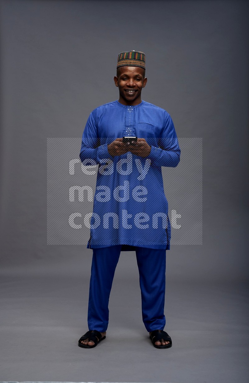 Man wearing Nigerian outfit standing texting on phone on gray background