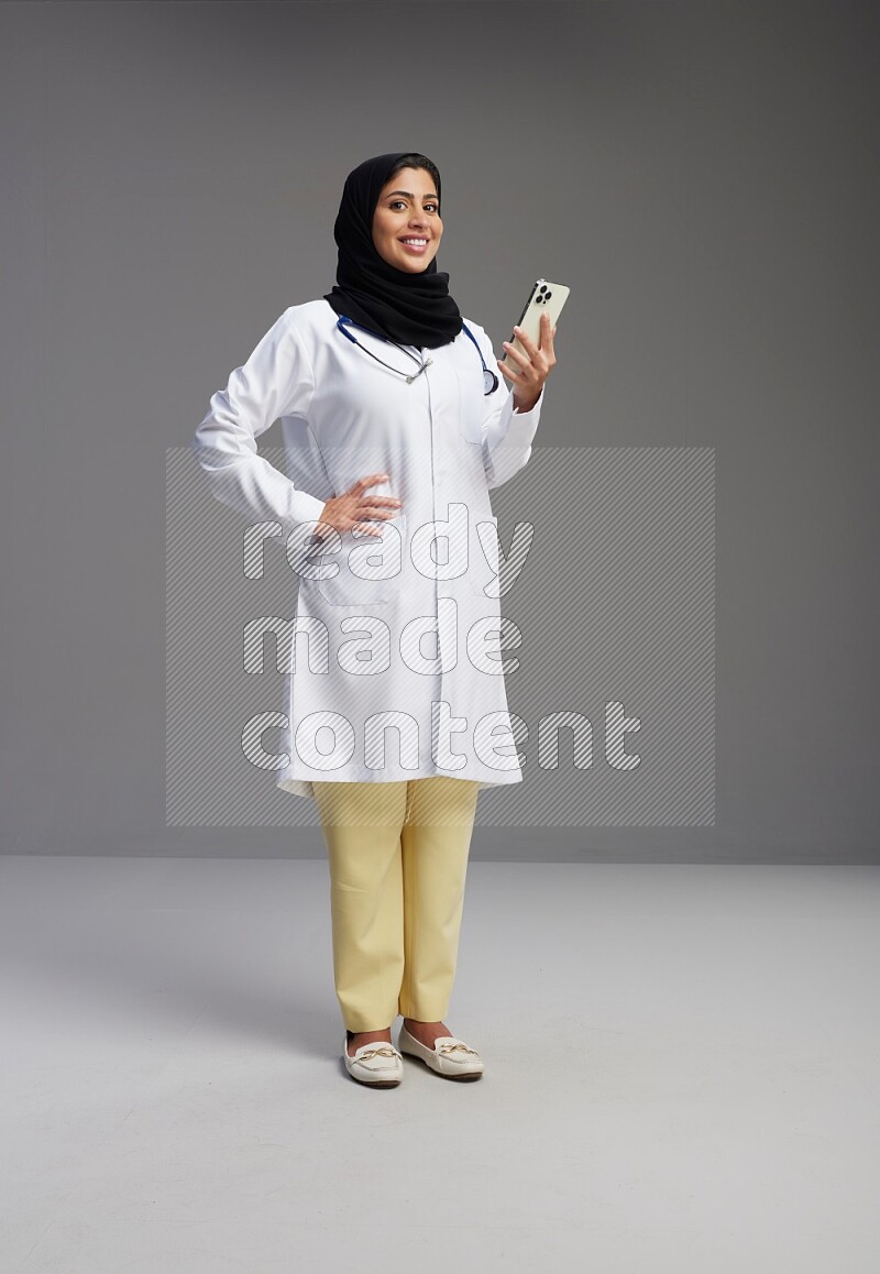 Saudi woman wearing lab coat with stethoscope standing texting on phone on Gray background