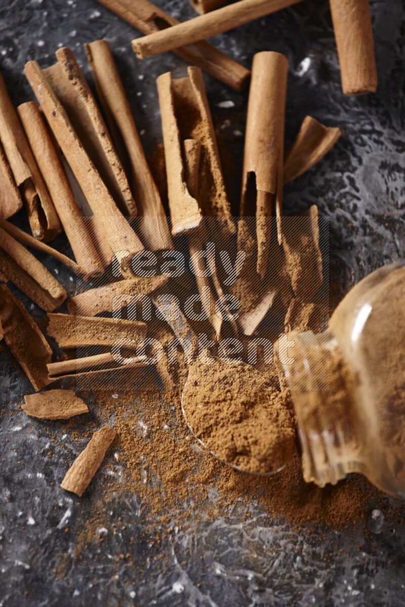 Herbal glass jar full cinnamon powder flipped and a metal spoon full of powder surrounded by cinnamon sticks on textured black background in different angles