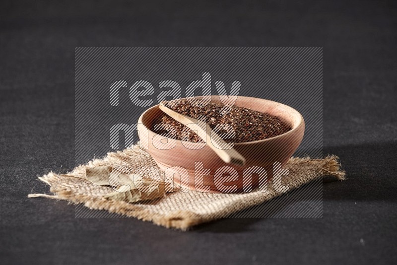 A wooden bowl full of flaxseeds with wooden spoon full of the seeds on it on burlap fabric on a black flooring