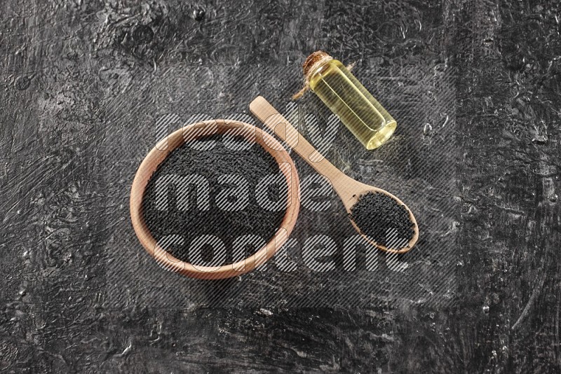 A wooden bowl and spoon full of black seeds and a bottle of black seeds oil on a textured black flooring