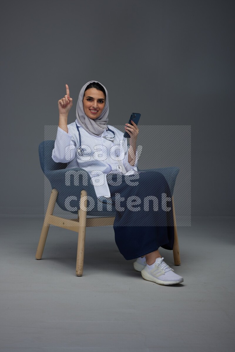 A doctor wearing a light gray head scarf setting on a dark grey chair and using a phone eye level on a grey background