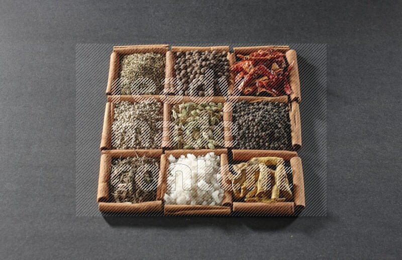 9 مربعات من أعواد القرفة ممتلئة بالهيل في المنتصف محاطة بأصابع الكركم والفلفل الأسود والفلفل الحار والبهار الحلو والكمون والفلفل الأبيض والريحان المجفف والملح على أرضية سوداء