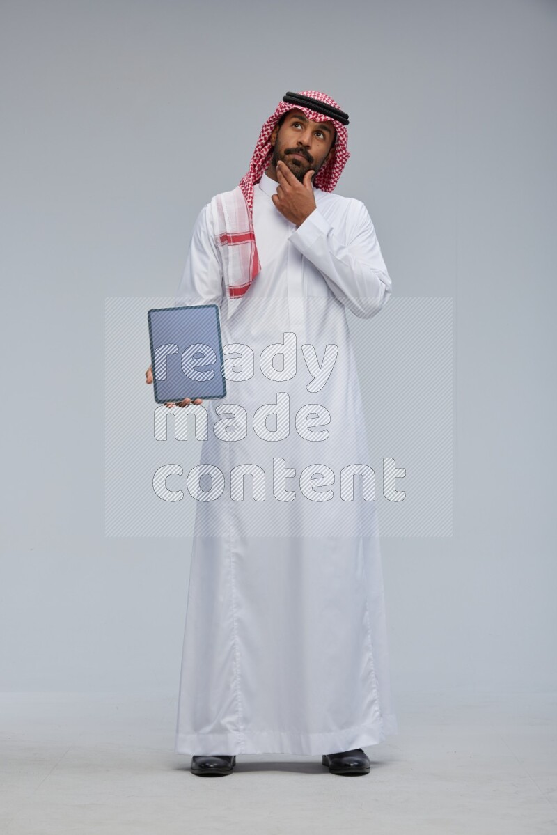 Saudi man Wearing Thob and shomag standing showing tablet to camera on Gray background
