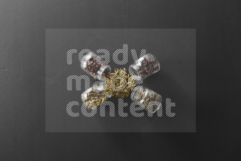 4 glass spice jars full of cloves, cardamom, basil and star anise on black flooring