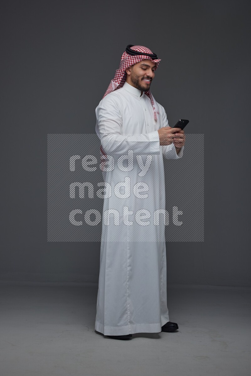 A Saudi man wearing Thobe and Shmagh standing and using his phone eye level on a gray background