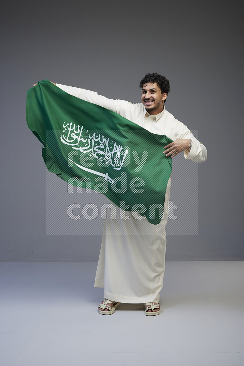 A Saudi man standing wearing thob holding big Saudi flag on gray background