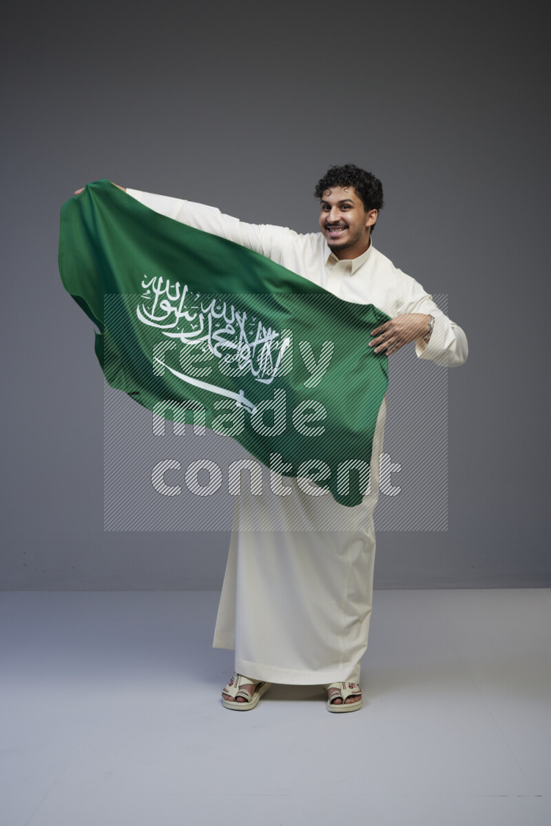 A Saudi man standing wearing thob holding big Saudi flag on gray background