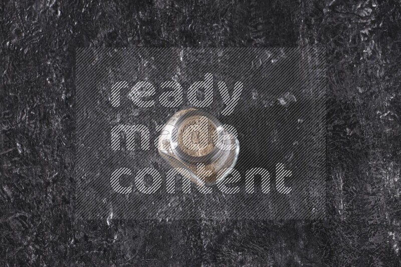 A glass spice jar full of black pepper powder on textured black flooring