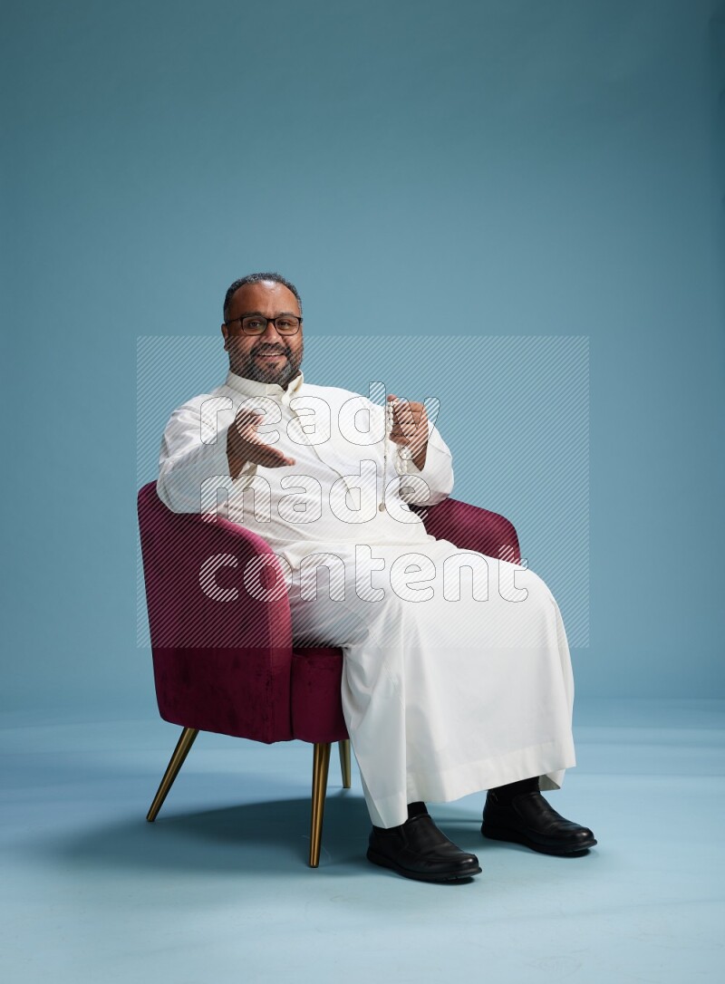 Saudi Man without shimag sitting on chair Interacting with the camera on blue background