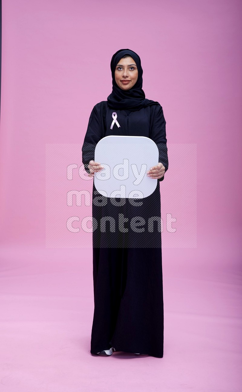 Saudi woman wearing pink ribbon on Abaya standing holding social media signs on pink background