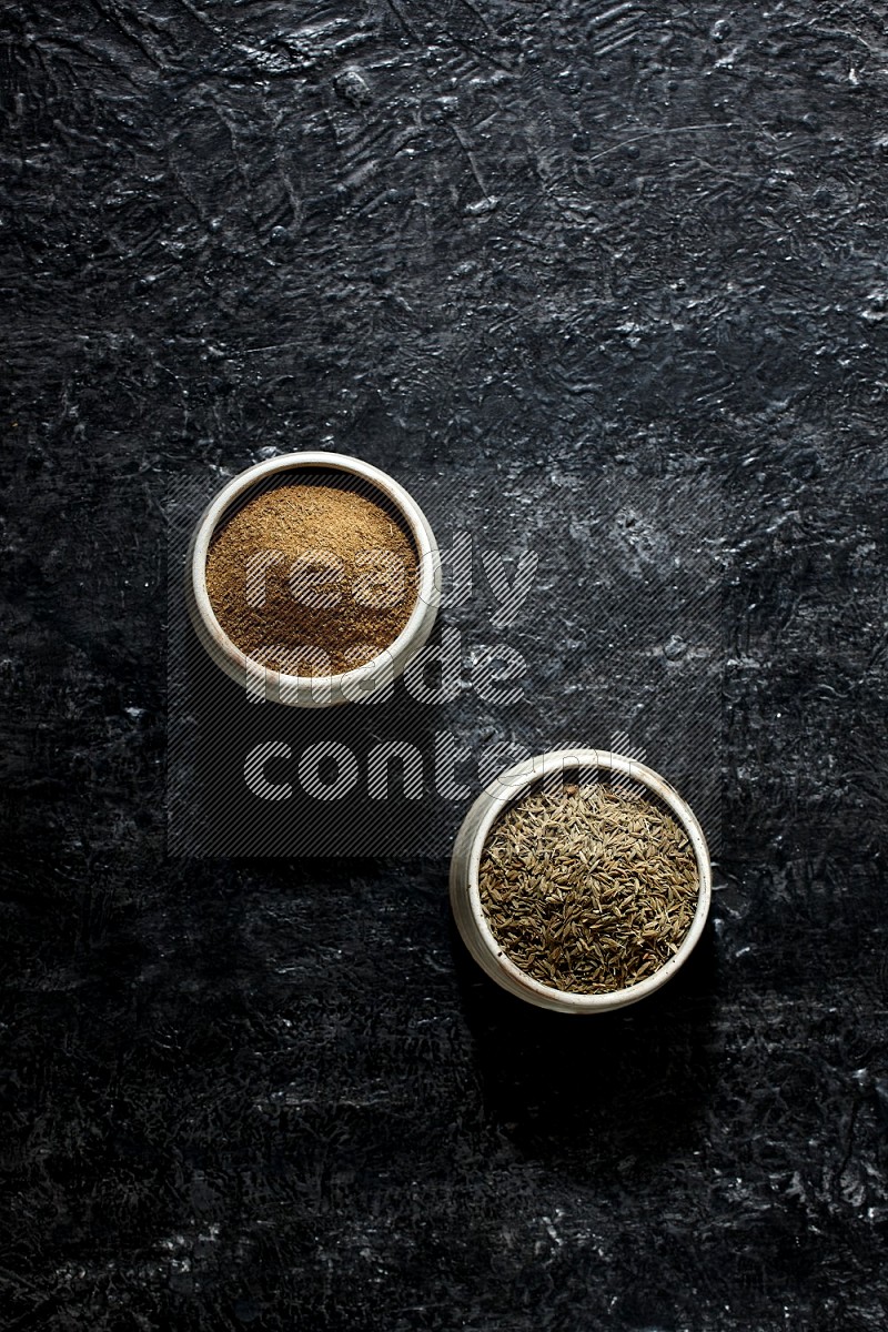 2 beige bowls, one full of cumin powder and the other full of seeds on a textured black flooring