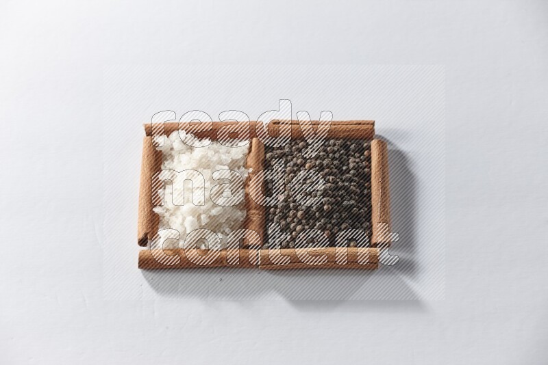 مربعان من أعواد القرفة ممتلئان بحبوب الفلفل الأسود والملح الأبيض علي خلفية بيضاء
