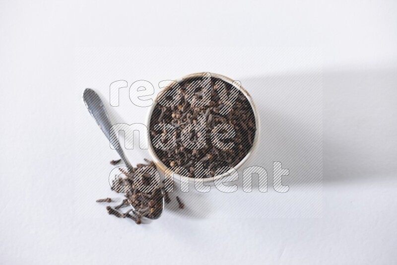 A beige ceramic bowl full of cloves and a metal spoon next to it on a white flooring