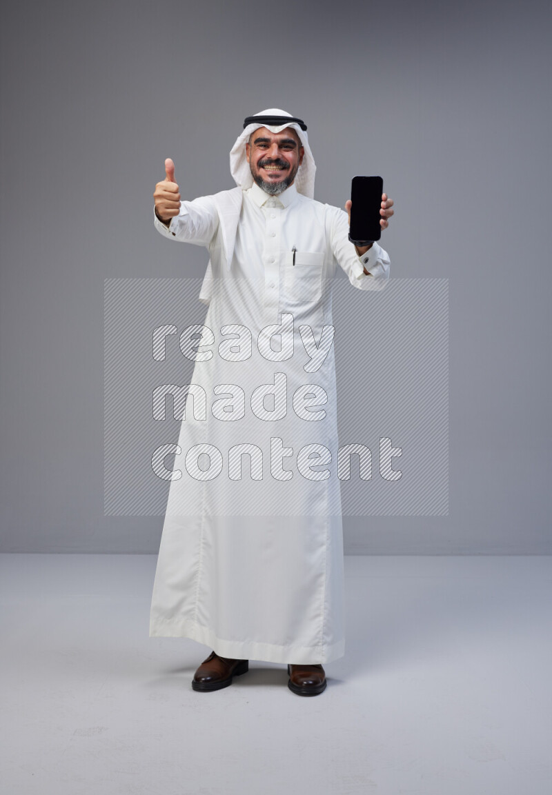 Saudi man Wearing Thob and white Shomag standing showing phone to camera on Gray background