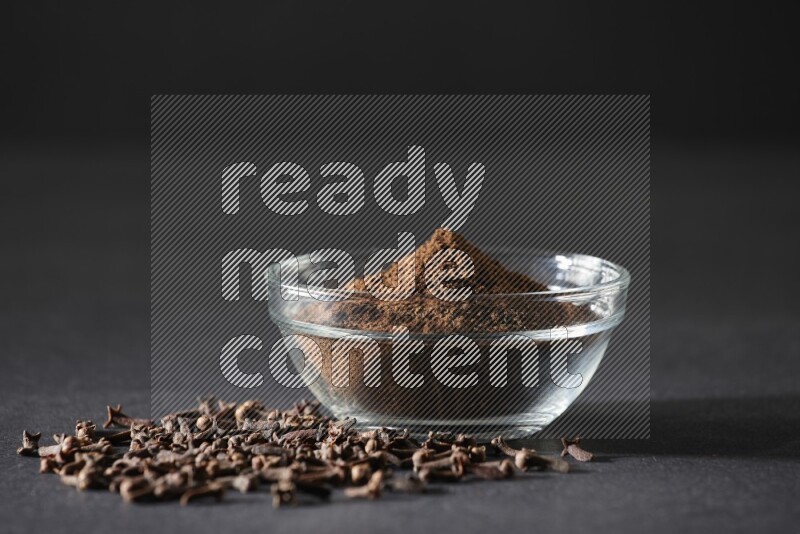 وعاء زجاجي ممتلئ ببودرة القرنفل مع حبوب القرنفل المتناثرة على أرضية سوداء