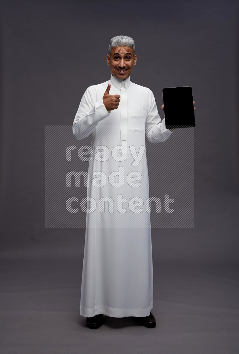 Saudi man wearing thob standing showing tablet to camera on gray background