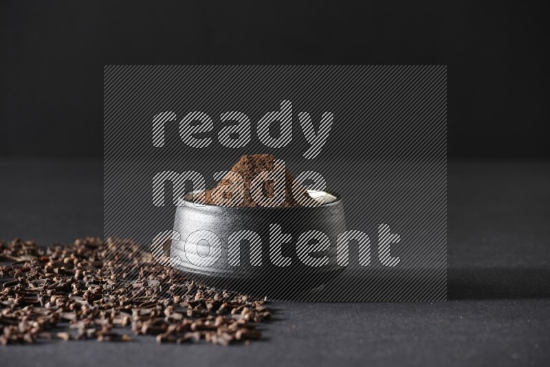 A black pottery bowl full of cloves powder and cloves whole grains on a black flooring