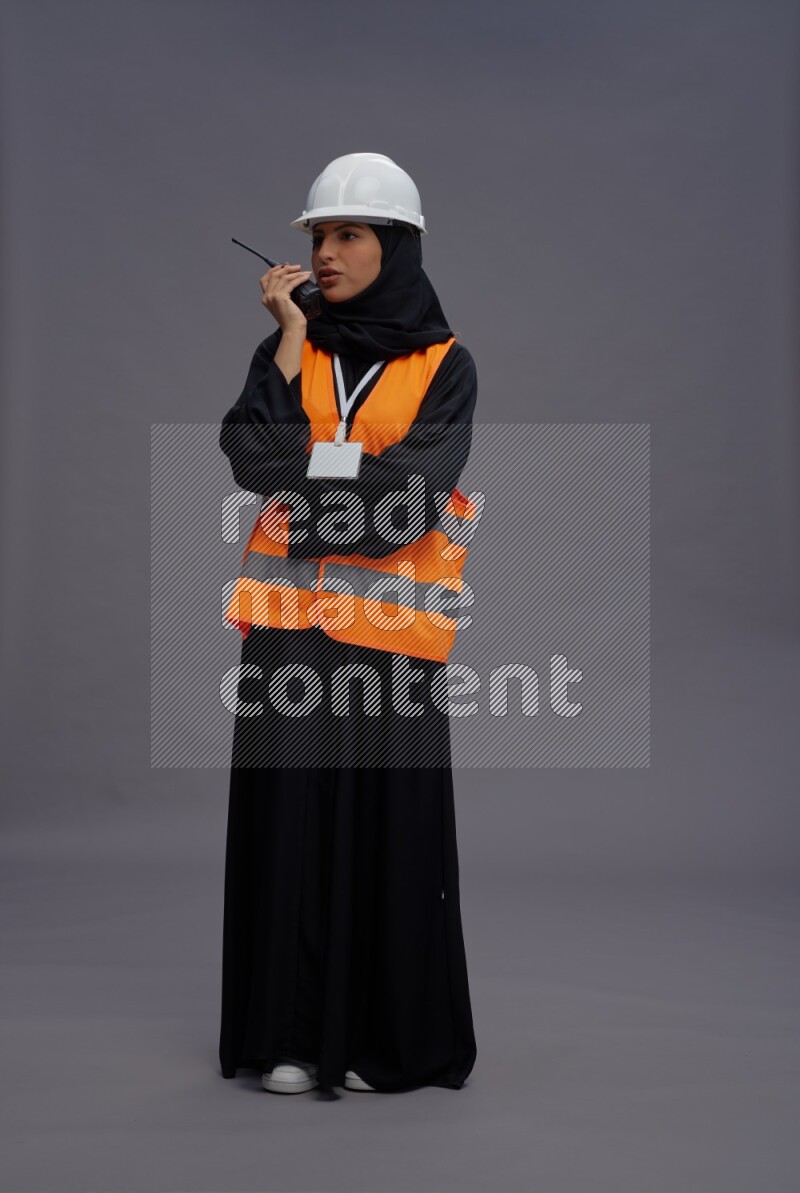 Saudi woman wearing Abaya with engineer vest with neck strap employee badge standing holding walkie-talkie on gray background