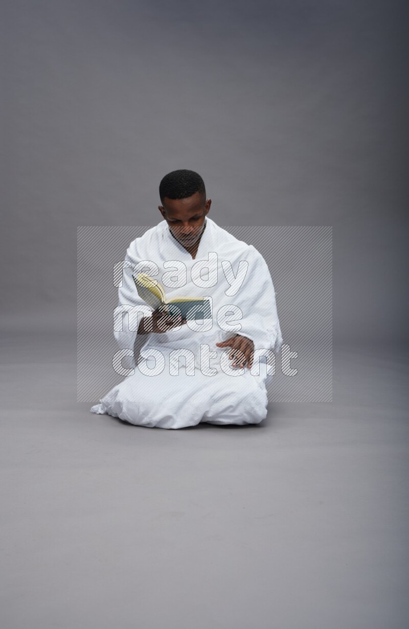 A man wearing Ehram sitting on floor reading quran on gray background