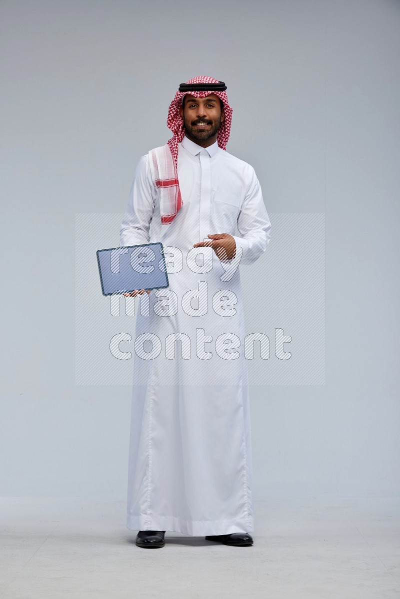 Saudi man Wearing Thob and shomag standing showing tablet to camera on Gray background