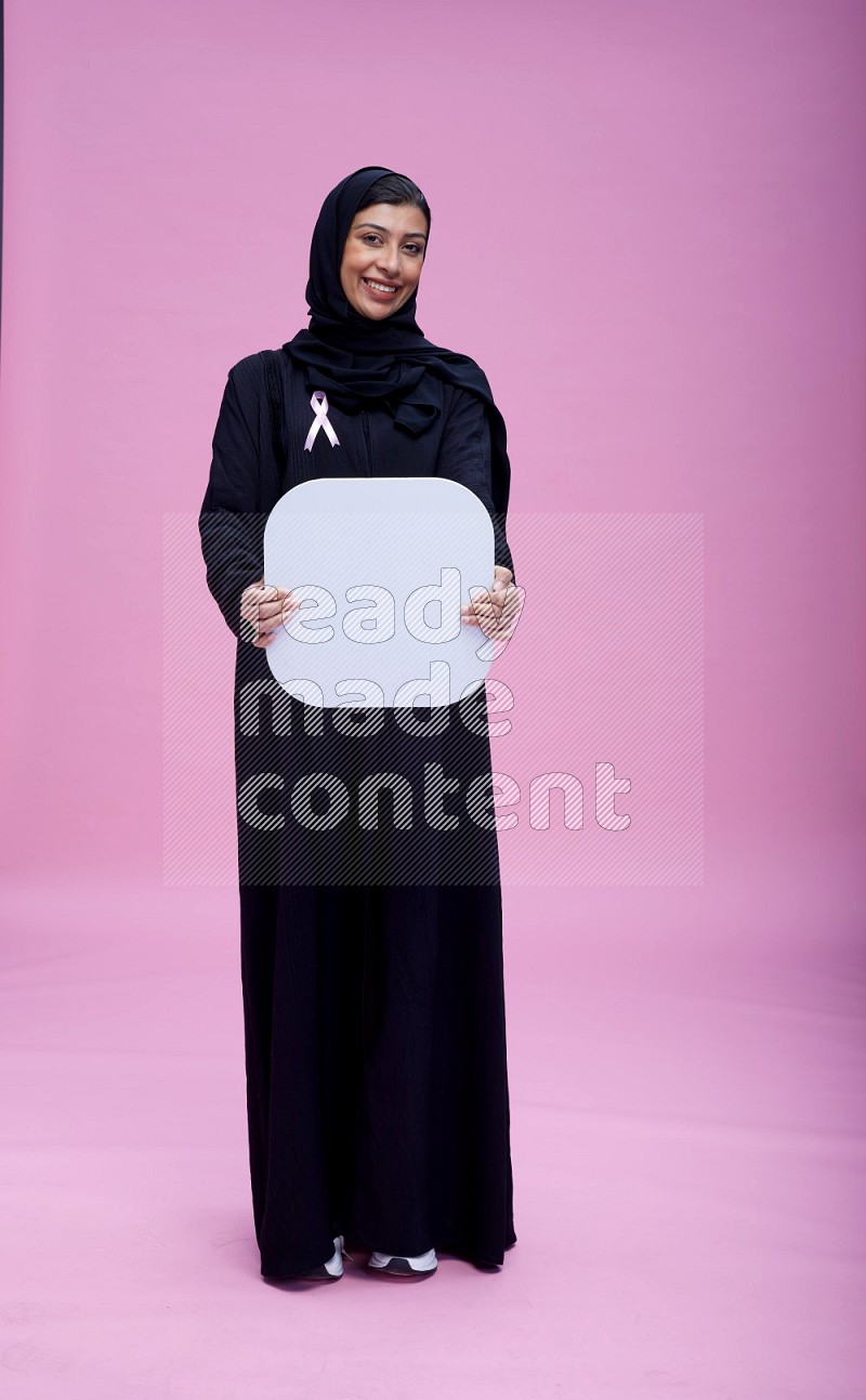 Saudi woman wearing pink ribbon on Abaya standing holding social media signs on pink background