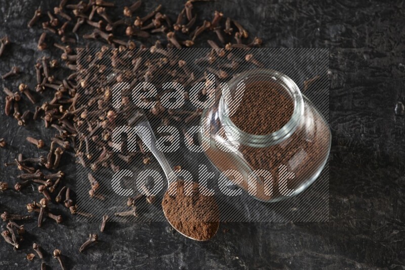 A glass spice jar and a metal spoon full of cloves powder with cloves spread on textured black flooring