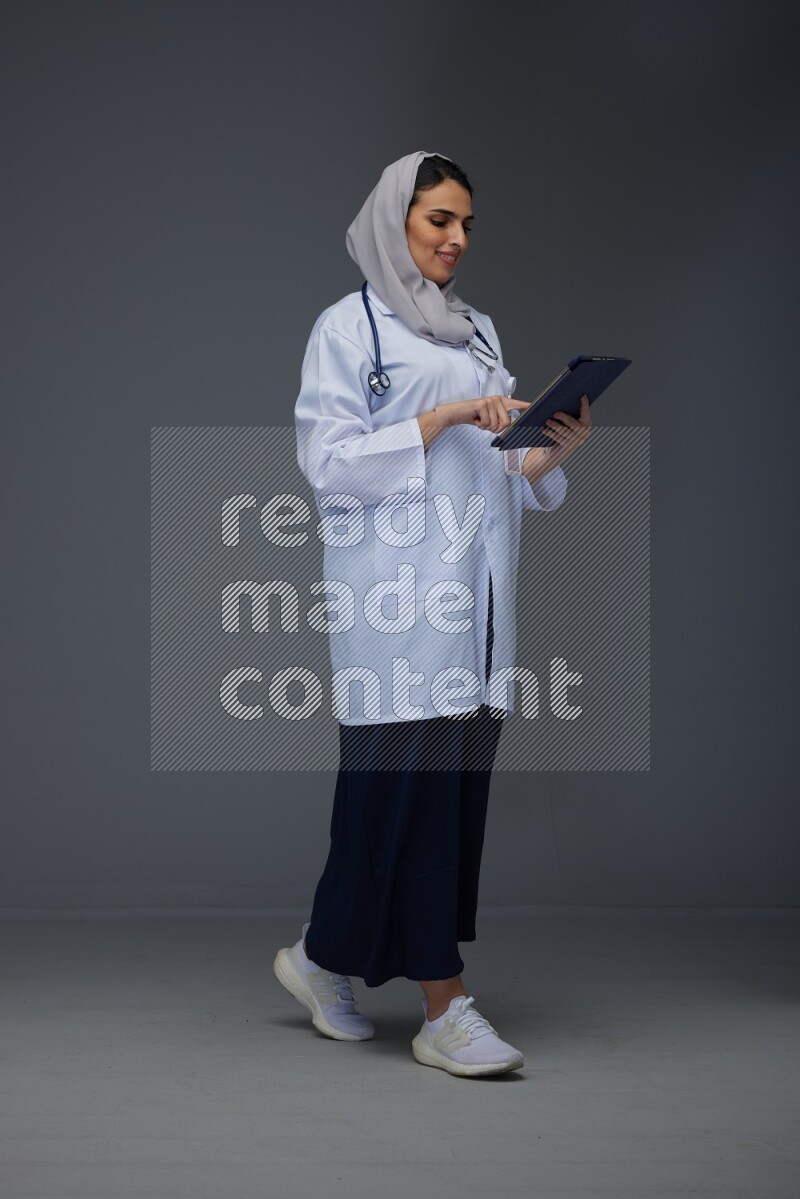 A Saudi doctor wearing a light gray head scarf standing and crossing her hands eye level on a grey background